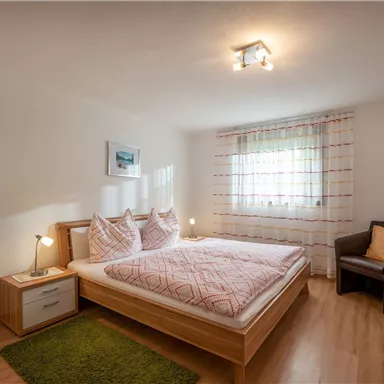 Ein gemütliches Schlafzimmer mit einem Doppelbett und hellen Textilien. Es gibt ein Fenster mit Vorhängen und einen bequemen Sessel daneben.