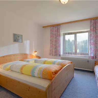 Ein helles Schlafzimmer mit zwei Betten und einem zusätzlichen Schlafsofa. Große Fenster lassen viel Tageslicht herein und die Wände sind mit Blumenvorhängen dekoriert.