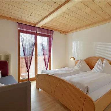 A cozy bedroom with wooden furniture and two single beds. The room has large windows with curtains and a quiet atmosphere.