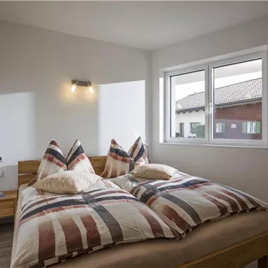 A cozy bedroom with a double bed and checked bedcovers. The window offers a view outside to a quiet residential area.