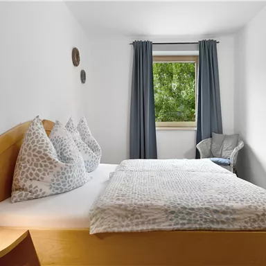 Ein helles Schlafzimmer mit einem Doppelbett und gemütlichen Kissen. Durch das Fenster ist viel Tageslicht und eine grüne Aussicht zu sehen.
