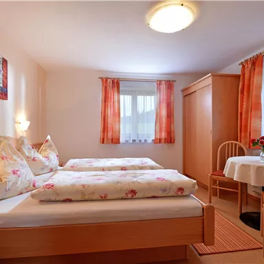 Ein gemütliches Zimmer mit zwei Einzelbetten und einem kleinen Tisch. Helle Vorhänge und Blumendekorationen schaffen eine einladende Atmosphäre.