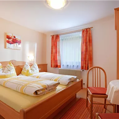 Ein gemütliches Schlafzimmer mit einem Doppelbett und hellen Vorhängen. Ein Tisch mit einer Blumenvase ergänzt die angenehme Atmosphäre.