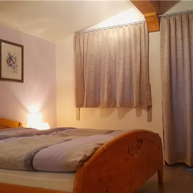 A cozy bedroom with a wooden bed and a soft duvet. The room is softly lit and has curtains at a window.