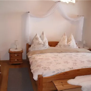 Ein gemütliches Schlafzimmer mit einem Holzbett und zwei Kissen in Herzform. Die Wände sind hell, und es gibt einen Kleiderschrank aus Holz sowie eine angenehme Beleuchtung.