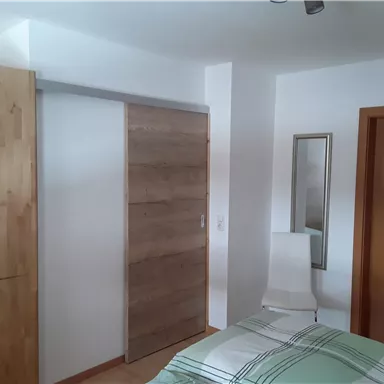 A bright bedroom with a bed and a wooden wardrobe. On the wall, there is a mirror and a light chair.