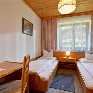 A bright room with two single beds and a wooden desk. The room has large windows and a cozy atmosphere.