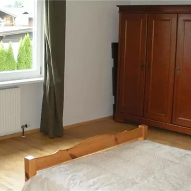 A simple bedroom with a wooden wardrobe and a large window. The floor is wooden and there is a bed with a plain duvet.