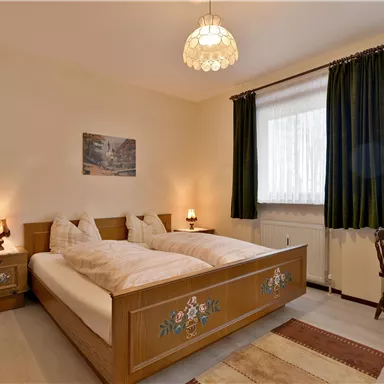 Ein gemütliches Schlafzimmer mit zwei Betten, Holzmöbeln und einem kleinen Tisch. Durch die Fenster fällt viel Tageslicht, und die Wände sind in neutralen Farben gehalten.