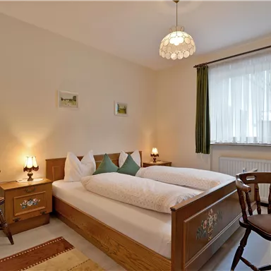 Ein gemütliches Schlafzimmer mit einem Doppelbett und Holzstuhl. Große Fenster mit Vorhängen sorgen für Tageslicht und eine einladende Atmosphäre.