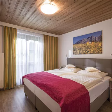 Ein gemütliches Schlafzimmer mit einem Doppelbett und modernen Möbeln. Große Fenster lassen viel Licht herein, und ein Bild mit einer Landschaft schmückt die Wand.