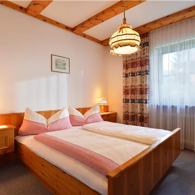 Ein gemütliches Schlafzimmer mit einem Doppelbett und Holzpaneelen. Es gibt eine charmante Lampe und einen hellen Vorhang, der Tageslicht hereinlässt.