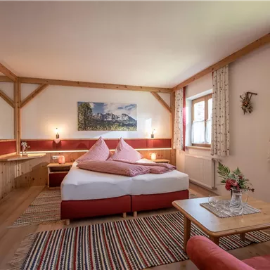 A cozy bedroom with a large bed, red pillows, and a television. The room has wooden details and large windows that provide plenty of daylight.