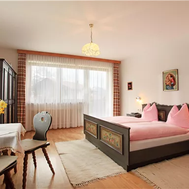 Ein gemütliches Schlafzimmer mit einem großen Doppelbett und rosa Kissen. Dazu gehören ein Tisch, Stühle und große Fenster mit viel natürlichem Licht.