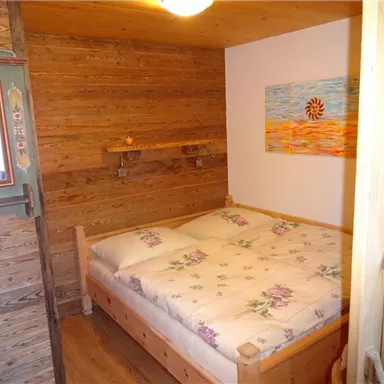 Ein gemütliches Schlafzimmer mit einem Holzrahmenbett und blühenden Kissen. Die Wände sind aus Holz, und an einer Wand hängt ein farbenfrohes Bild.