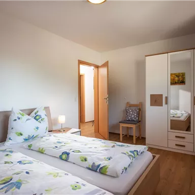 A bright bedroom with two beds and fresh flower-patterned bedding. The room has a wardrobe and a chair.