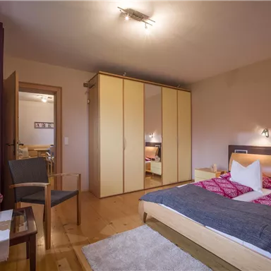 A cozy bedroom with a large bed and a wardrobe. The room is bright and inviting, with a chair and a small table.