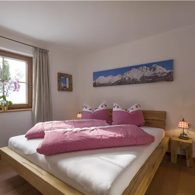 A cozy bedroom with a large bed and pink bedding. The window offers a view of beautiful plants and a mountain landscape on the wall.