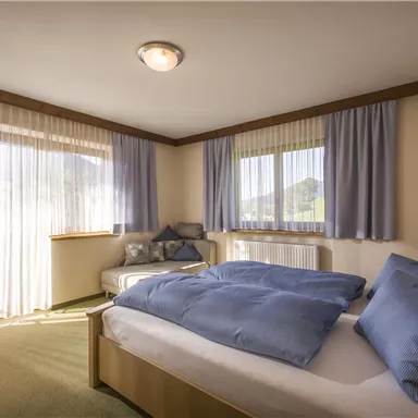A cozy bedroom with a large bed and blue pillows. Bright curtains let in daylight and offer a view of the surrounding landscape.