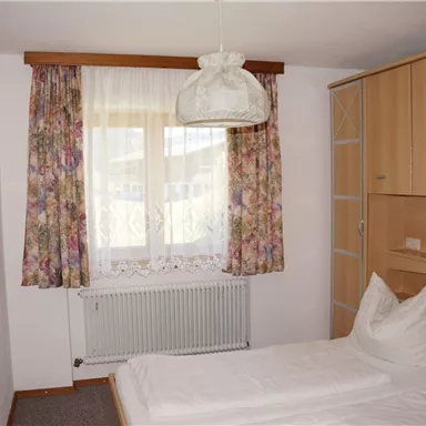 Ein einfaches, helles Schlafzimmer mit zwei Betten und einem großen Fenster. Vorhänge und eine Lampe verleihen dem Raum eine gemütliche Atmosphäre.