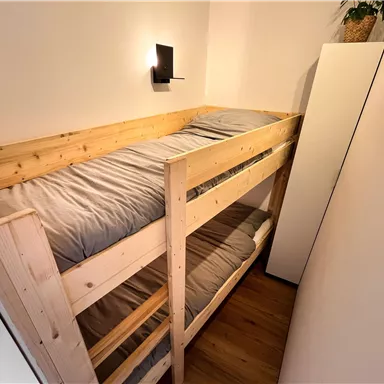 Ein einfaches Etagenbett aus Holz mit grauen Bettlaken. Der Raum ist minimalistisch und hell eingerichtet.