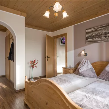 A cozy bedroom with a wooden double bed and soft colors. On the wall hangs a picture of a landscape, and there is a small flower arrangement on the nightstand.
