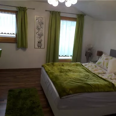 Ein gemütliches Schlafzimmer mit einem großen Bett und grüner Bettwäsche. Grüne Vorhänge und ein kleiner Schreibtisch schaffen eine einladende Atmosphäre.