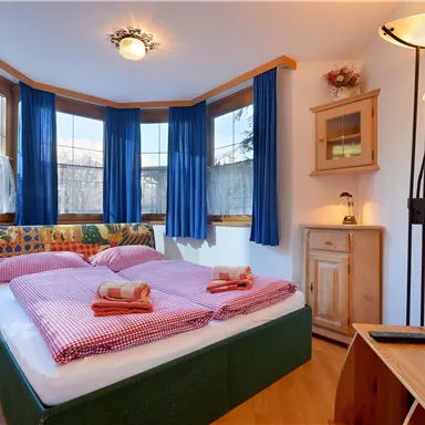 A cozy bedroom with a large double bed and colorful pillows. Large windows with blue curtains provide plenty of light.