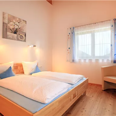 Ein gemütliches Schlafzimmer mit einem Doppelbett und sanfter Beleuchtung. Das Zimmer hat ein Fenster mit Vorhängen und einen bequemen Stuhl in der Ecke.