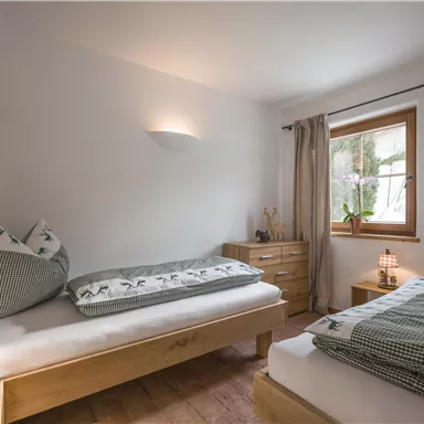 A cozy bedroom with two single beds and natural light. The decor is simple and modern, with wood and textile elements.