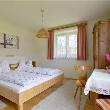 A cozy bedroom with two beds and a table. The room has large windows that let in plenty of daylight.