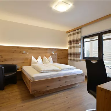 Ein modernes Zimmer mit Holzmöbeln und einem großen Fenster. Es gibt ein Doppelbett, einen Schreibtisch und einen gemütlichen Sessel.