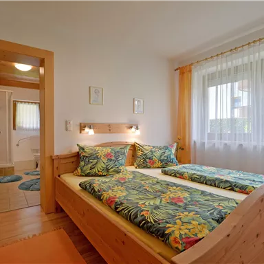 Ein gemütliches Schlafzimmer mit einem Doppelbett und bunten Bettbezügen. Die großen Fenster lassen viel Licht herein und bieten einen Blick nach draußen.