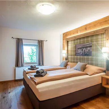 A cozy bedroom with two beds and warm wooden furniture. Large windows provide plenty of daylight.