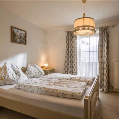 A cozy bedroom with a double bed and two nightstands. Large windows with curtains let in plenty of light.
