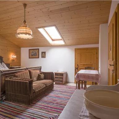 A cozy, bright room with a wooden ceiling and a sun-drenched window. It is furnished with a sofa, a table, and a classic rug.