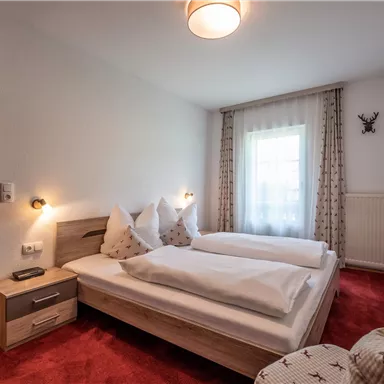 Ein gemütliches Schlafzimmer mit zwei Einzelbetten und roten Teppichen. Große Fenster sorgen für viel Tageslicht und eine einladende Atmosphäre.