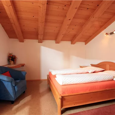 A cozy bedroom with a wooden carved ceiling. There is a double bed, a blue armchair, and a bedside table.