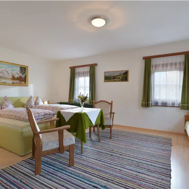 Ein gemütliches Zimmer mit zwei Betten und einem Tisch. Große Fenster lassen viel Licht herein und das Farbenspiel schafft eine einladende Atmosphäre.