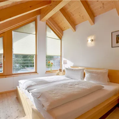 Ein gemütliches Schlafzimmer mit einem Holzinterieur und Dachschräge. Große Fenster lassen viel Tageslicht herein und bieten einen Blick ins Freie.