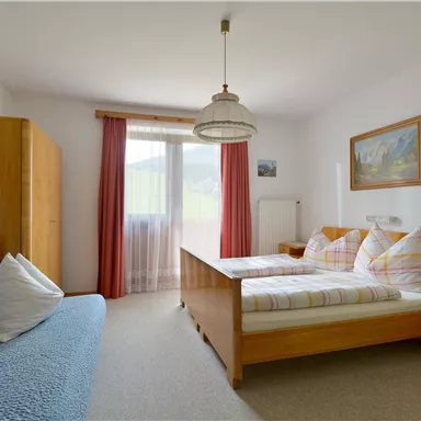 A bright bedroom with two beds and a sofa. Large windows with natural light and a warm, inviting decor.