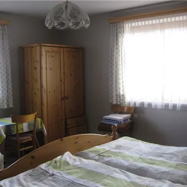 Ein einfaches Schlafzimmer mit zwei Einzelbetten und einem Holzschrank. Das Zimmer hat große Fenster mit Vorhängen und einen kleinen Tisch mit einem Stuhl.