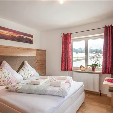 A cozy bedroom with a large bed and colorful pillows. Large windows with red checkered curtains and a small dining table create an inviting atmosphere.