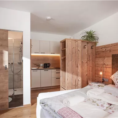 A cozy bedroom with wooden decor and a bathroom area. The amenities include a bed, a small desk, and a kitchen.