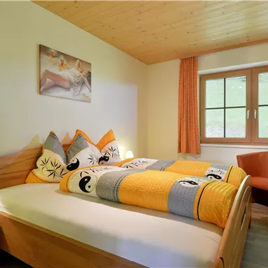 Ein gemütliches Schlafzimmer mit einem Doppelbett und bunten Kissen. Das Zimmer hat große Fenster und warmes Holzdekor.