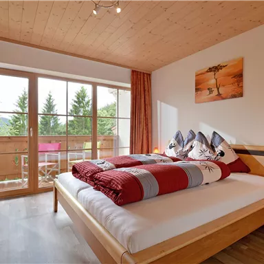 Ein gemütliches Schlafzimmer mit einem großen Bett und bunten Decken. Der Raum hat große Fenster mit Blick auf einen Balkon und grüne Bäume.