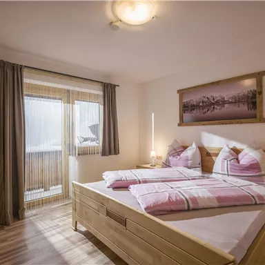 A cozy bedroom with two beds and wooden furniture. Large windows provide plenty of daylight and a view of the balcony.