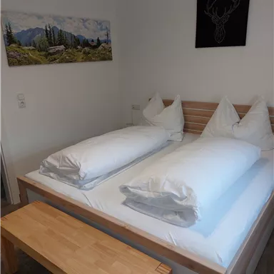 Ein gemütliches Schlafzimmer mit einem Doppelbett, zwei Kissen und einer Holzbank. An der Wand hängt ein Landschaftsbild sowie ein Kunstwerk mit einem Hirschmotiv.