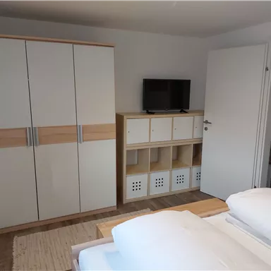 Ein einfaches Schlafzimmer mit zwei Betten, einem Schrank und einem Fernseher. An der Wand steht ein Regal mit Stauraum.