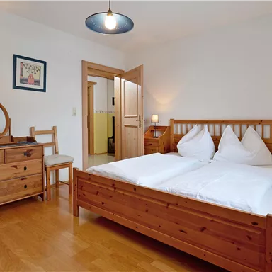 A cozy bedroom with wooden furniture and a double bed. The walls are painted light and there are some decorative pictures.